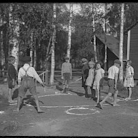 Tar Pot: First, a circle was drawn on the ground. Smaller circles were then drawn around the main circle, at an equal distance from each other, all of them touching the main circle. There had to be one smaller circle less than there were participants in the game.   One of the players was elected “bear”. The others went to stand in the smaller circles, facing each other. The bear then ran around the circle, behind the back of the others, carrying a stick in his hand. At some point, he would drop the stick behind the back of one of the other players, trying not to attract too much attention to it. The idea, however, was for the other player to notice the stick as soon as possible, pick it up and start running around the circle, in the opposite direction as the bear.   Now there was an empty circle in the field, and the bear and the other player, now carrying the stick, raced to see which one of them could reach the empty circle first. The one who finished second, got to be bear for the next round. If the bear was able to complete his round without the other one noticing the stick in the first place, the latter one, then, would become bear. (Vilhelm Slotte, 14 years old, Nedervetil.) The picture is taken in the village of Nedervetil in 1932.   Originator: Maximilian Stejskal.  Archive collection: The Society of Swedish Literature in Finland (SLS), sls.finna.fi SLS 407 b, 39