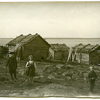 Three children in a fishing village. Sheds with roofs of water reed and a fisherman’s cabin with a birch-bark roof. 1900–1920’s.  Photographer: unknown.  Archive collection: The Society of Swedish Literature in Finland (SLS), sls.finna.fi ÖTA 247, 164