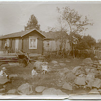 The fishing village of Skatan in Kristinestad. Two cabins and a rack for drying fishing nets. In the foreground three small children in their Sunday best. Outside one of the cabins is a woman. The picture also shows a container for salt and a barrel. 1900–1930’s.  Photographer: unknown.  Archive collection: The Society of Swedish Literature in Finland (SLS), sls.finna.fi ÖTA 247, 174